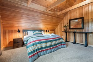 Quarto com teto de madeira, cama com roupa de cama listrada, mesa de cabeceira, cômoda, espelho e uma pequena mesa com uma planta.