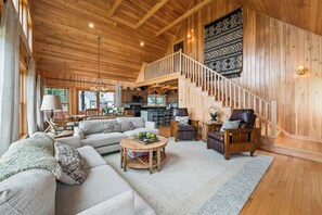 Uma sala de estar espaçosa com teto alto, paredes de madeira, uma escada e uma cozinha ao fundo.