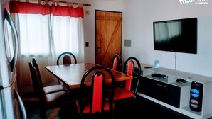 Un coin repas avec une table en bois et des chaises à revêtement rouge, un téléviseur fixé au mur et une porte en bois.