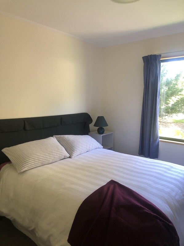 A neatly made bed with white and striped bedding, a dark headboard, a bedside lamp, and a window with curtains.