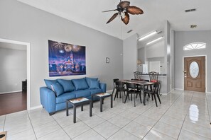 A living room with a blue sofa, a coffee table, and a dining area with a table and chairs. - Awesome 3BR/2BA Home, Private Pool (Extra Daily Charge to Heat), Disney Theme! (Kissimmee)
