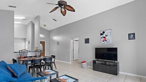 Un salon moderne avec un canapé bleu, une table à manger avec des chaises, un ventilateur de plafond, un téléviseur à écran plat et des œuvres d’art accrochées aux murs.