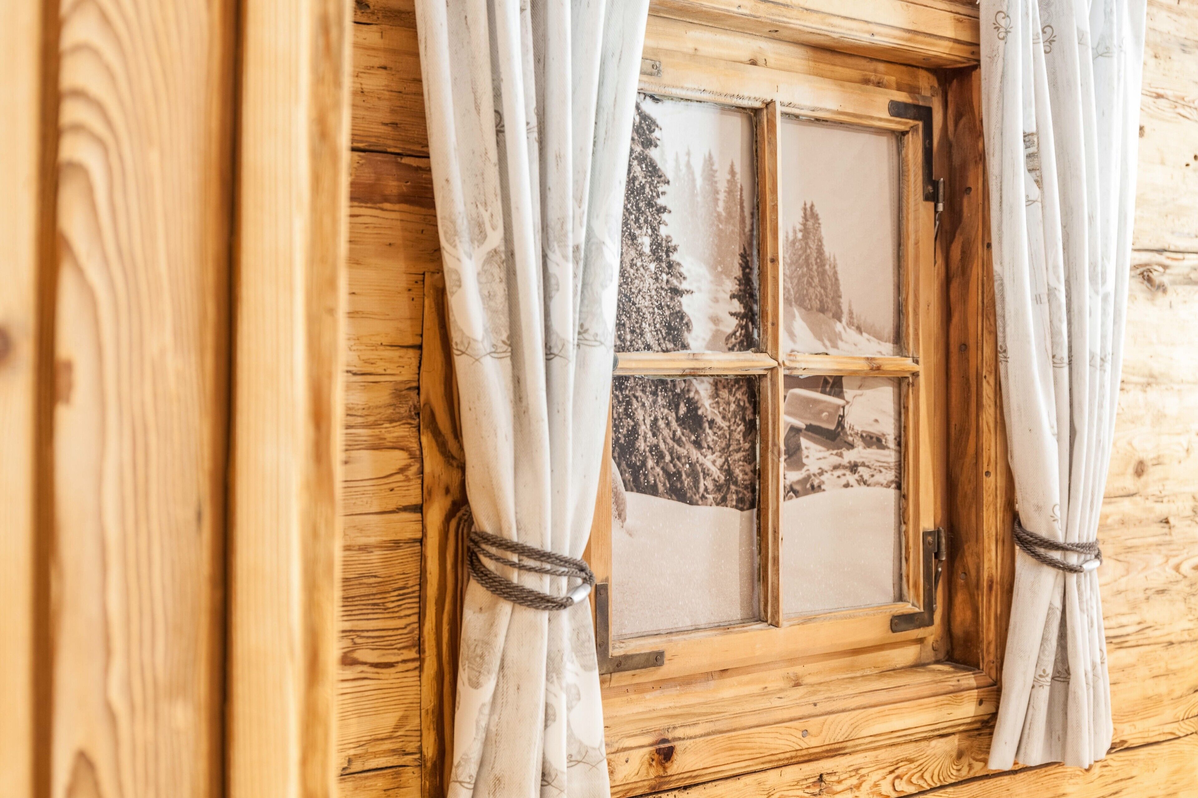 Una finestra di una baita in legno con tende ripiegate, che mostra una vista panoramica su un paesaggio innevato.