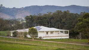 Una casa bianca con tetto spiovente e pannelli solari, immersa in prati verdi e alberi, con colline sullo sfondo.