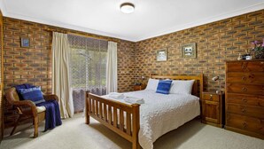 Une chambre avec un mur décoré d’un motif en briques, un lit en bois, une chaise en osier, une commode en bois et une fenêtre ornée de rideaux légers.