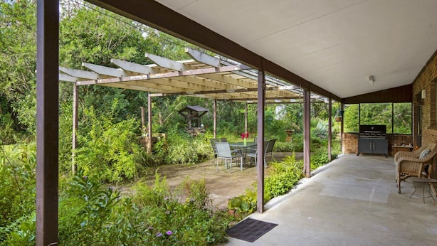 Une terrasse couverte, agrémentée d’une table et de chaises, entourée d’une végétation luxuriante et d’une structure en pierre.