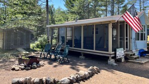 A small cabin with a porch, outdoor seating, and an American flag. - The Bears Den cabin w screened porch in campground on bike trail. (Boulder Junction)