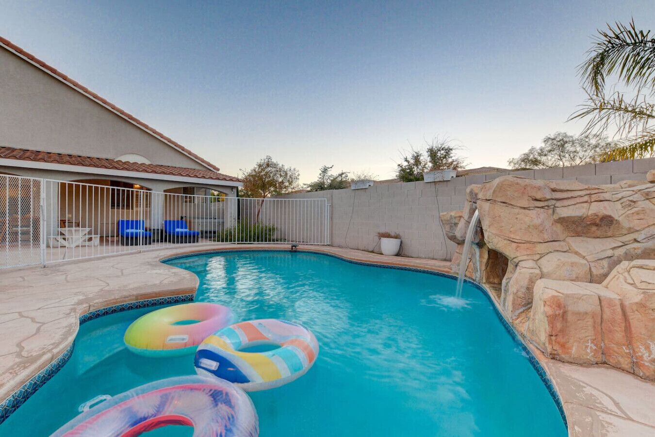 A swimming pool with a small waterfall feature, surrounded by a white fence and a stone wall.