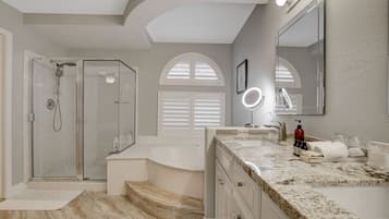 A bathroom with a shower, bathtub, vanity with a mirror, and a large window with shutters.