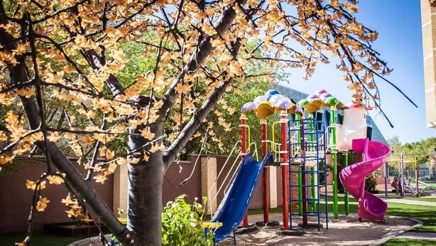 A playground with slides and climbing structures, surrounded by trees and a building in the background. - Europa Residence (Atyrau)