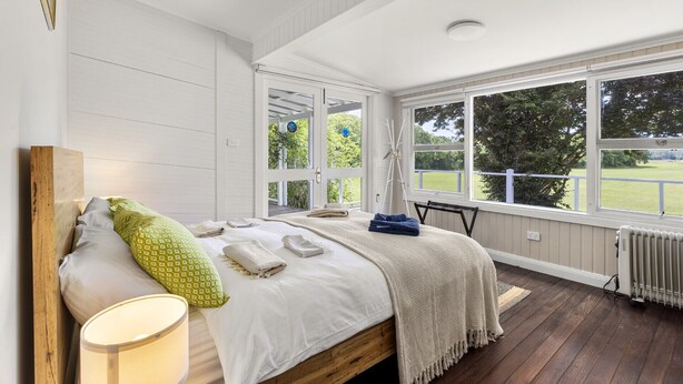 Ein Schlafzimmer mit Bett, Nachttisch, Lampe und Blick auf eine grüne Landschaft durch große Fenster.