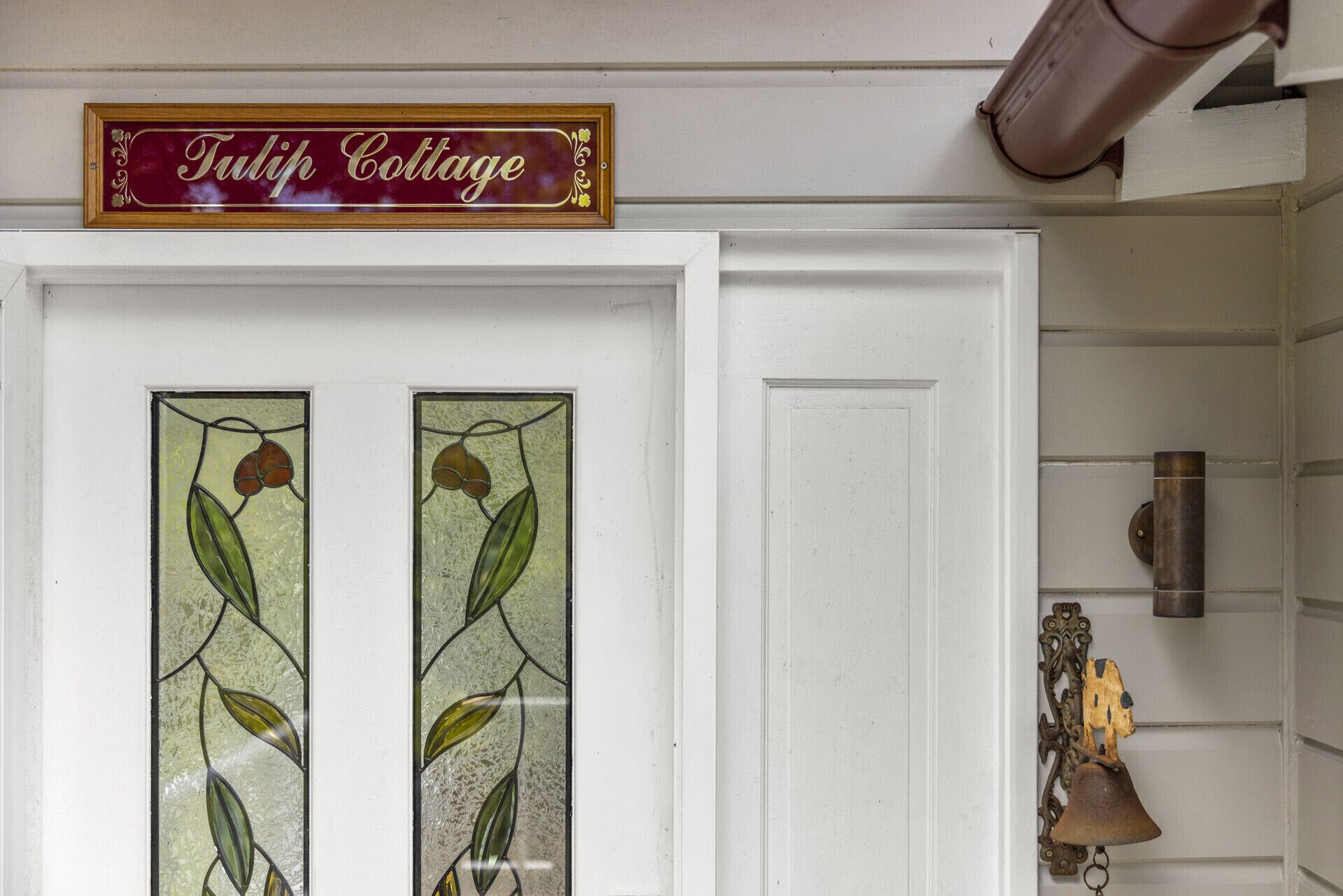Una puerta blanca con un vitral que muestra motivos de hojas. Sobre la puerta, un cartel dice 'Tulip Cottage'.
