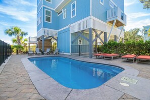 Zona de piscina con un edificio azul, mobiliario exterior y cielo despejado.