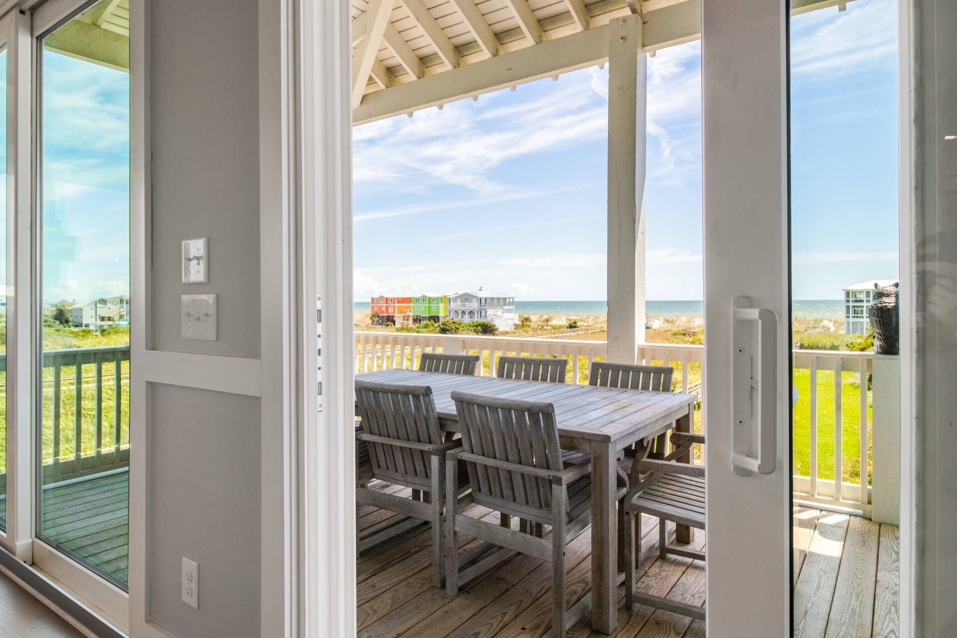 A covered outdoor area with a wooden table and chairs, overlooking a beach with colorful houses and a clear sky.