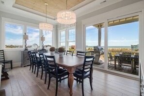 Un comedor con mesa y sillas de madera, ventanas amplias con vista a la playa y techo con vigas a la vista.