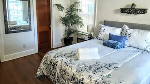 Un dormitorio con una cama grande, una planta en maceta, una mesita de noche con lámpara y un espejo en la pared.