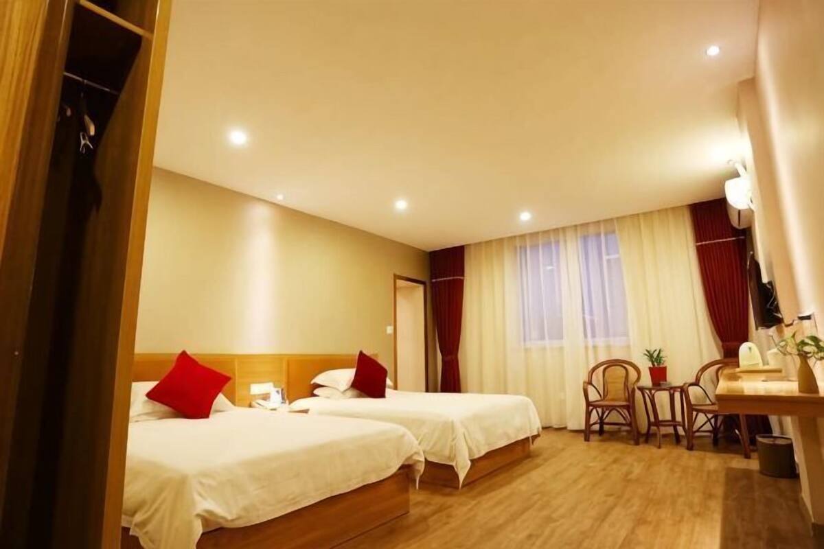 A hotel room with two beds, a desk, a chair, a small table, a potted plant, and a window with curtains.