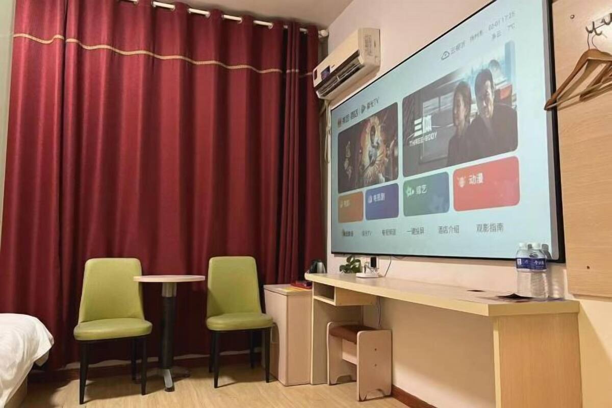 A hotel room with a bed, two green chairs, a desk with a computer, and a projector screen displaying movie options.
