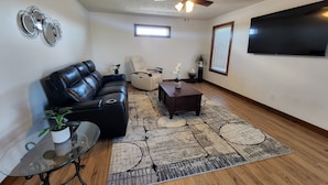 A living room with a black leather sofa, a glass coffee table, a beige armchair, a wooden coffee table, a patterned rug, a ceiling fan, a TV, and a small plant. - Perfect for a TEAM of people! (Hastings)