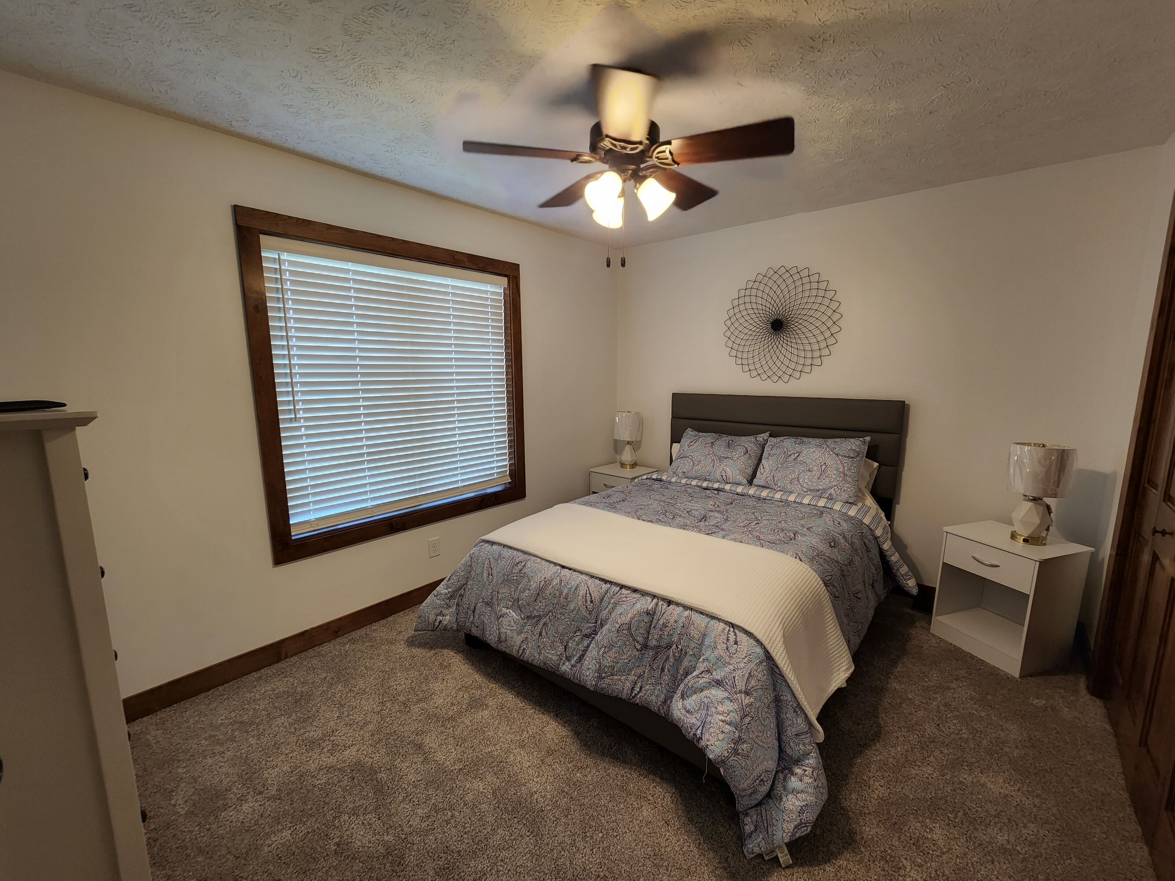 Un dormitorio con cama, ventilador de techo, decoración en la pared, mesita de noche y ventana con persianas.