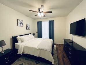 A bedroom with a bed, a ceiling fan, a TV, a dresser, and a window with curtains.