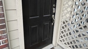 Una puerta negra con cerradura electrónica, un felpudo y una valla blanca con celosías.