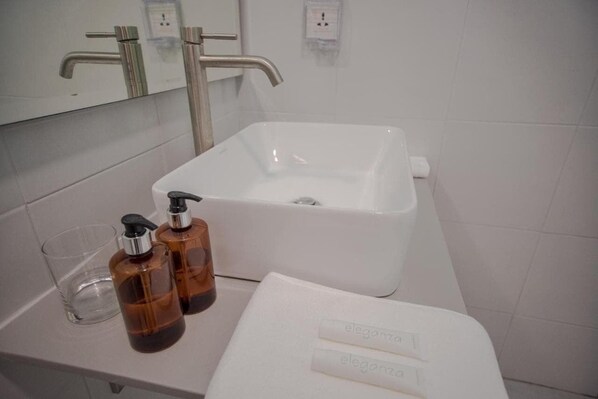 Et badeværelse med en hvid vask, to brune sæbedispensere, et klart glas og et hvidt håndklæde med 'eleganza' på.