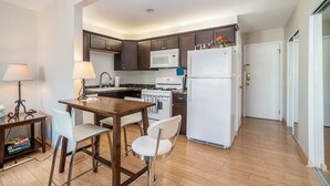 A compact kitchen with dark wooden cabinets, a white microwave, a stove, and a refrigerator. There is a dining table with two chairs and a small side table with a lamp. - Beautiful Pool and Lake View (South Haven)
