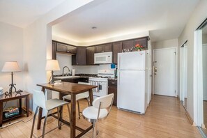 A compact kitchen with dark wooden cabinets, a white microwave, a stove, and a refrigerator. There is a dining table with two chairs and a small side table with a lamp.