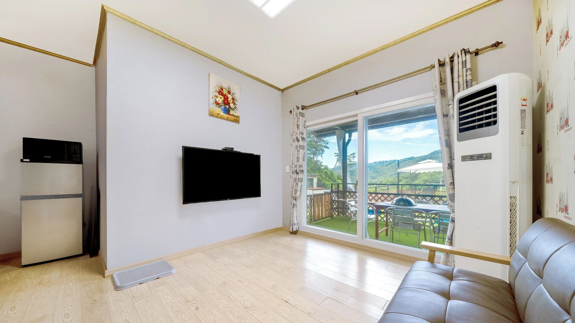 Una sala de estar con televisión de pantalla plana, un sofá, un refrigerador y un amplio ventanal con vista a un paisaje verde.