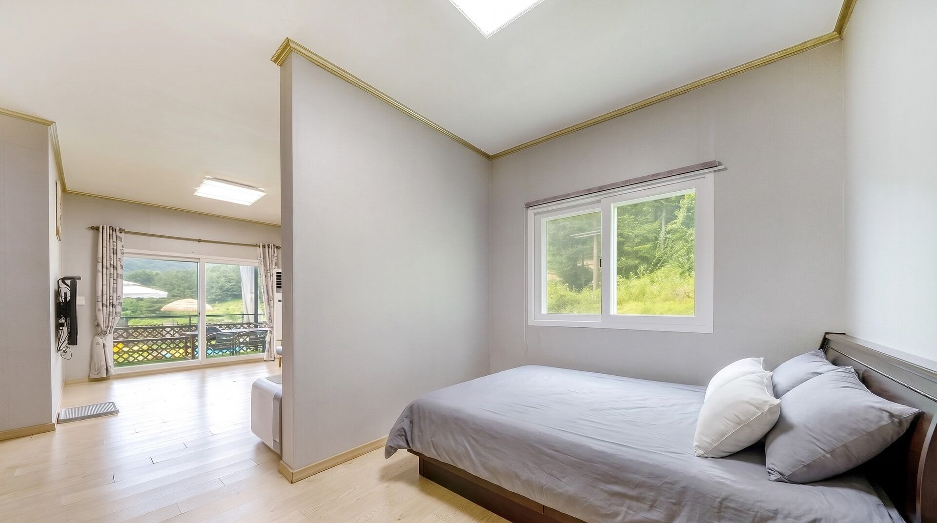 A bedroom with a bed, a window with a view of greenery, and a balcony with outdoor furniture.