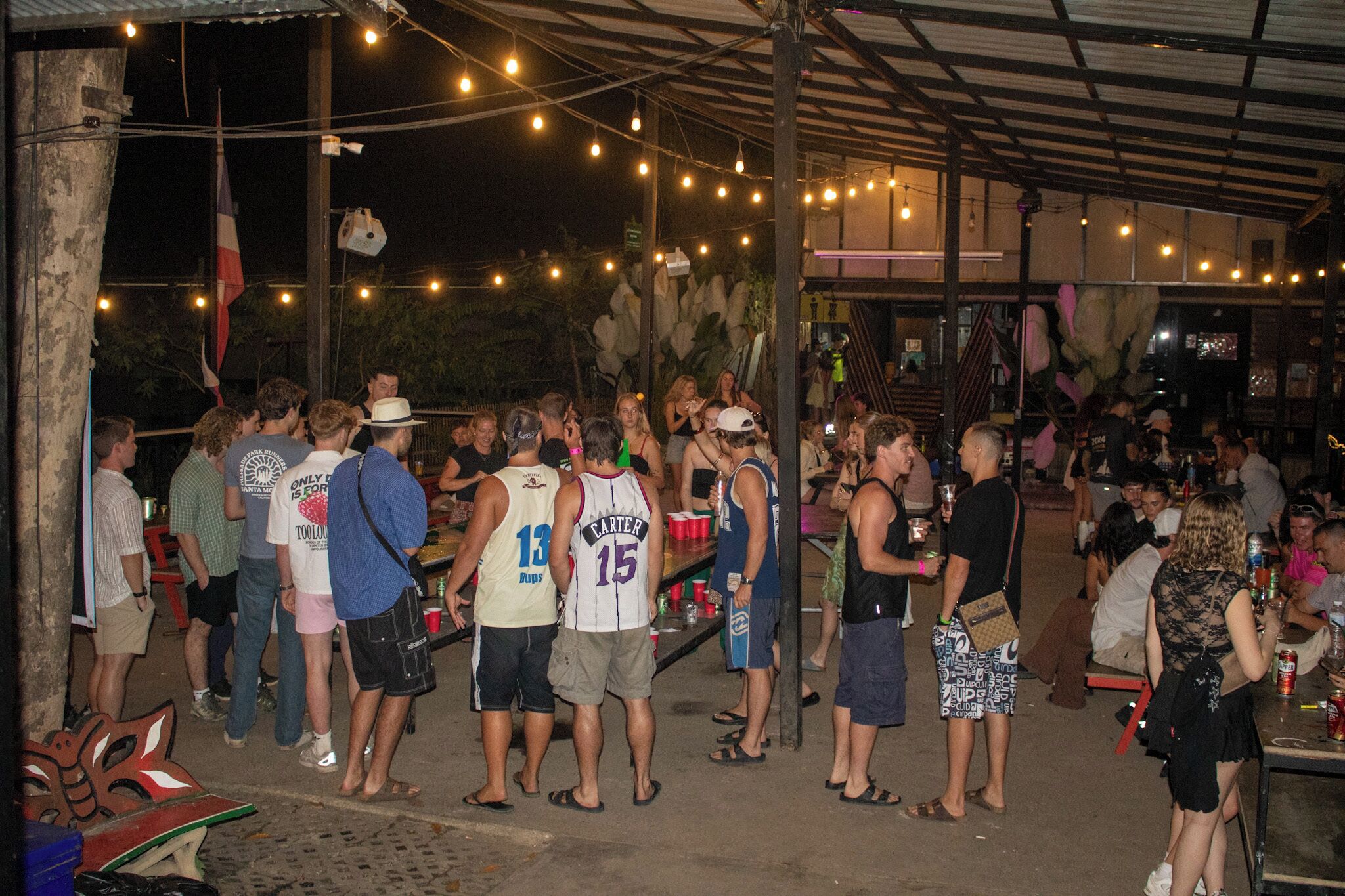 A crowded outdoor area with people standing and socializing, under a covered structure with string lights.