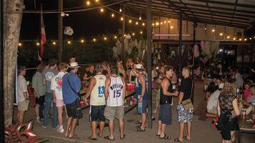A crowded outdoor area with people standing and socializing, under a covered structure with string lights.