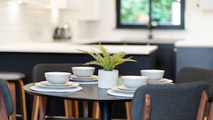 Un coin repas avec une table ronde dressée pour quatre personnes, entourée de chaises. Un pot de plante orne la table, et une cuisine moderne est visible au fond de la pièce.