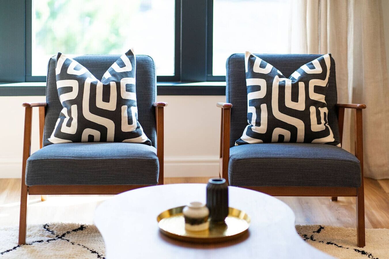 Two armchairs with patterned cushions, a small round table with a decorative item, and a window with curtains in the background.