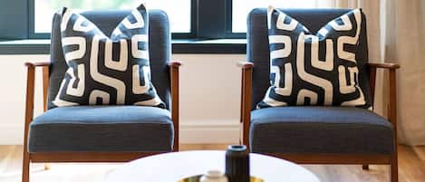 Dos sillones con cojines estampados, una mesita redonda con un objeto decorativo y una ventana con cortinas al fondo.