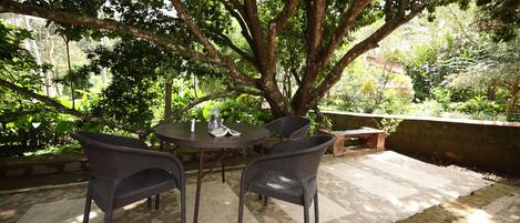 Une terrasse extérieure avec une table ronde et deux chaises en osier, entourée d’un sol carrelé et d’un grand arbre qui offre de l’ombre.