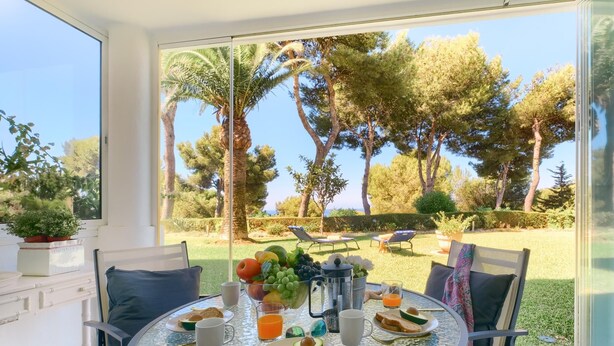 Una mesa de comedor con alimentos para el desayuno, vista a un jardín con árboles y césped, y mobiliario para disfrutar al aire libre.