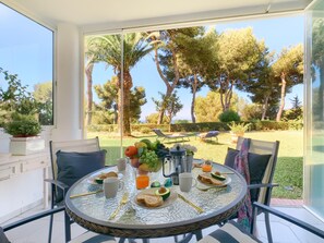 Une table à manger avec des aliments pour le petit-déjeuner, une vue sur un jardin avec des arbres et une pelouse, et des sièges d’extérieur.