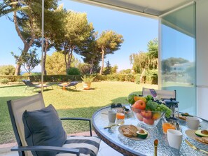 Un espace de restauration extérieur baigné de lumière, avec une table dressée pour le petit-déjeuner, donnant sur un jardin agrémenté d’arbres et d’une pelouse.