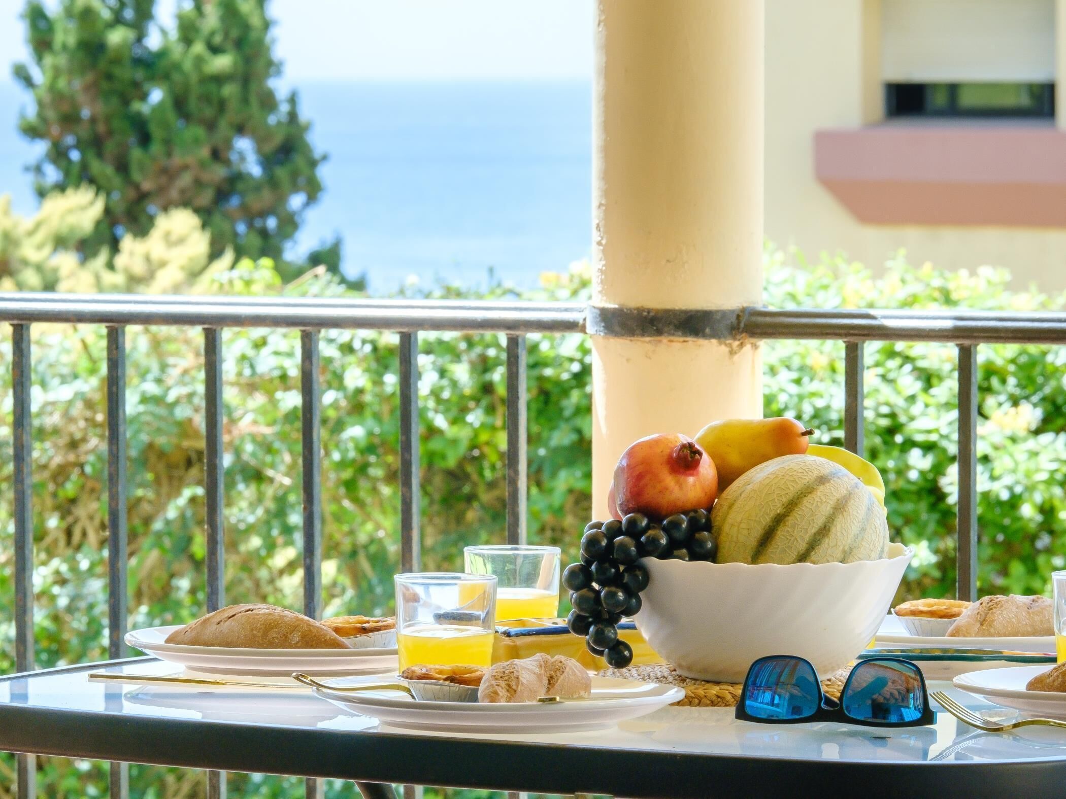 A breakfast setup on a balcony with a view of the sea, including fruits, pastries, and glasses of orange juice.