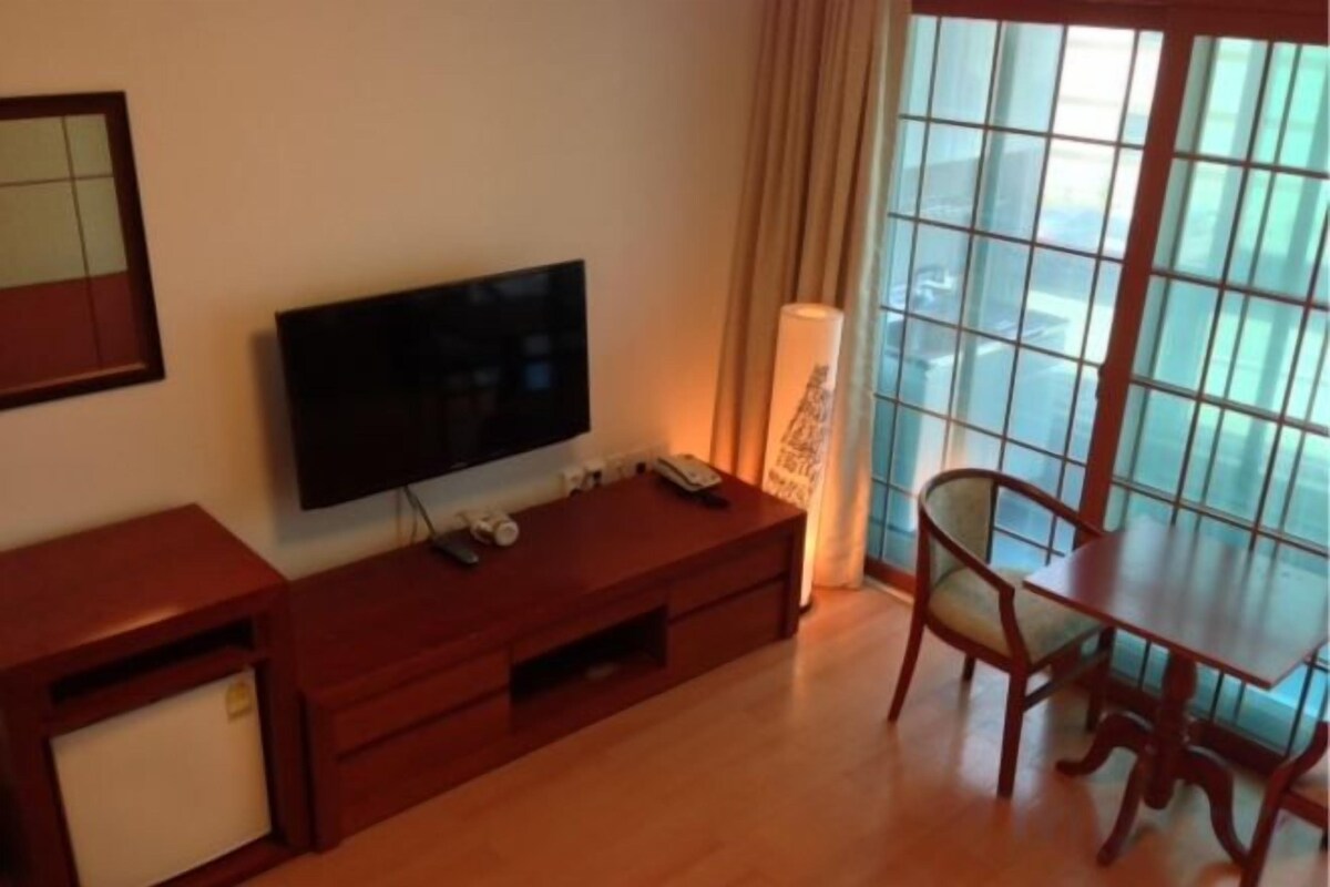 A room with a flat-screen TV on a wooden stand, a small table with a chair, and a large window with curtains.