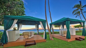 Två utomhusliggande paviljonger med sängar och sittplatser, med utsikt över en strand och havet.