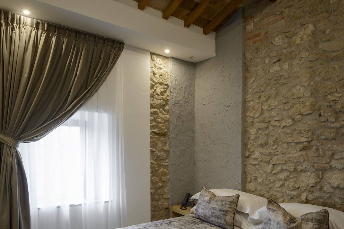 A bedroom with a stone wall, a wooden ceiling, a bed with a patterned bedspread, and a nightstand with a telephone.