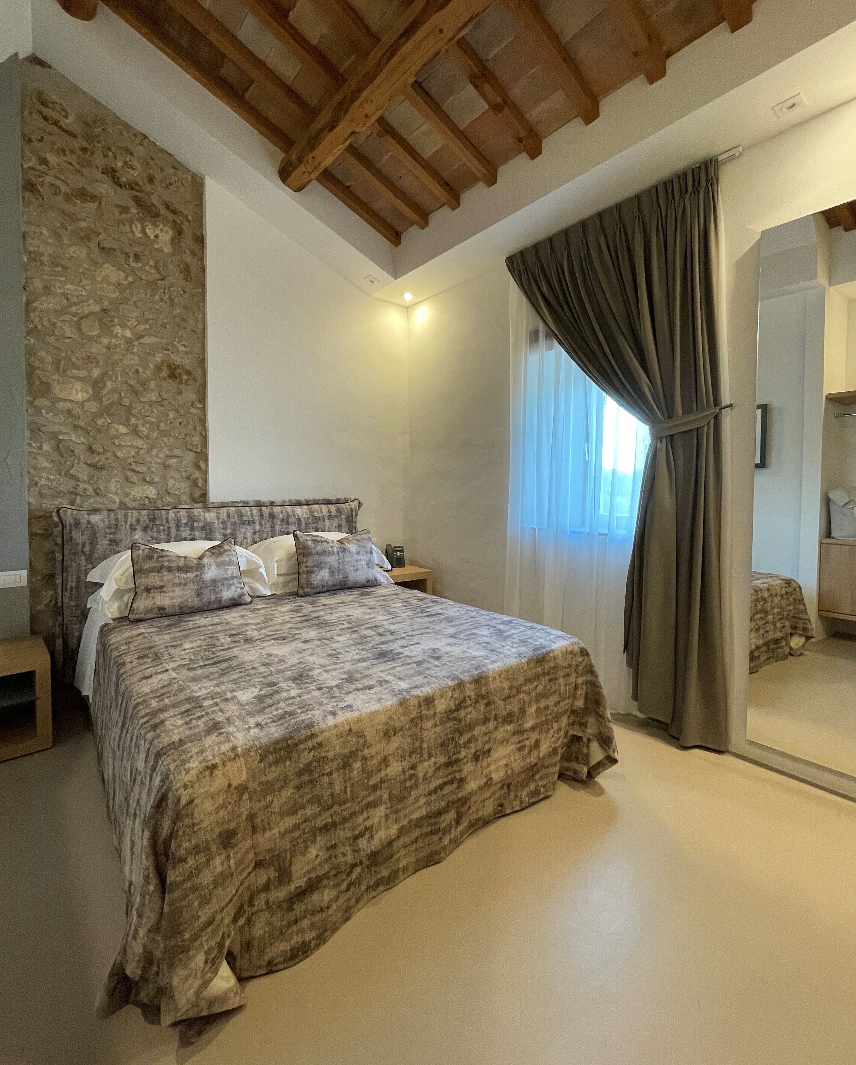 Ein Schlafzimmer mit einer Steinswand, einer Holzbalkendecke, einem Bett mit gemustertem Überwurf, einem Nachttisch und einem Fenster mit Vorhängen.
