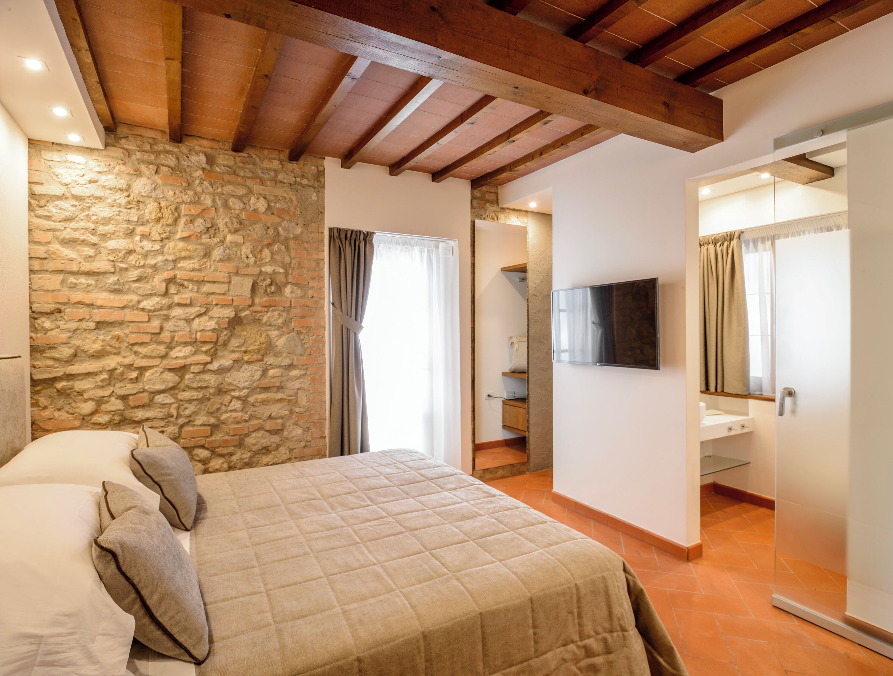A bedroom with a stone wall, wooden beams, a bed with pillows, a TV, and a bathroom with a mirror and shower.