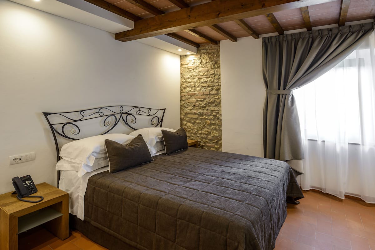 A bedroom with a bed, a nightstand with a phone, a stone wall, and a window with curtains.