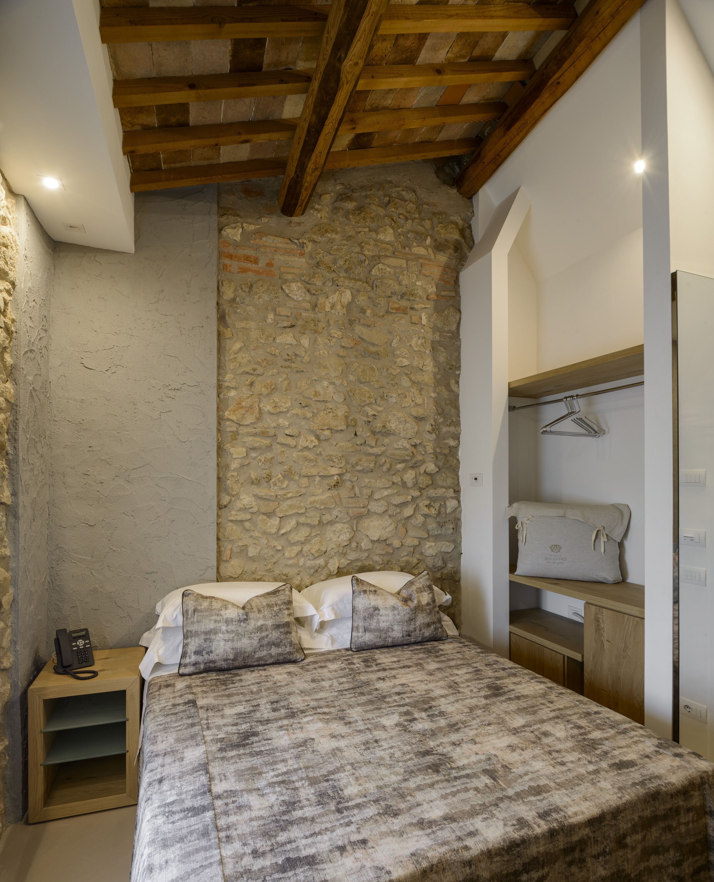 A bedroom with a stone wall, a wooden bed frame, a bedside table with a lamp, and a hanging towel rack.