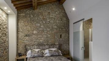 Une chambre avec un mur en pierre, un plafond en bois, un lit recouvert d’une couvre-lit à motifs, une table de chevet avec un téléphone et une armoire avec un miroir.