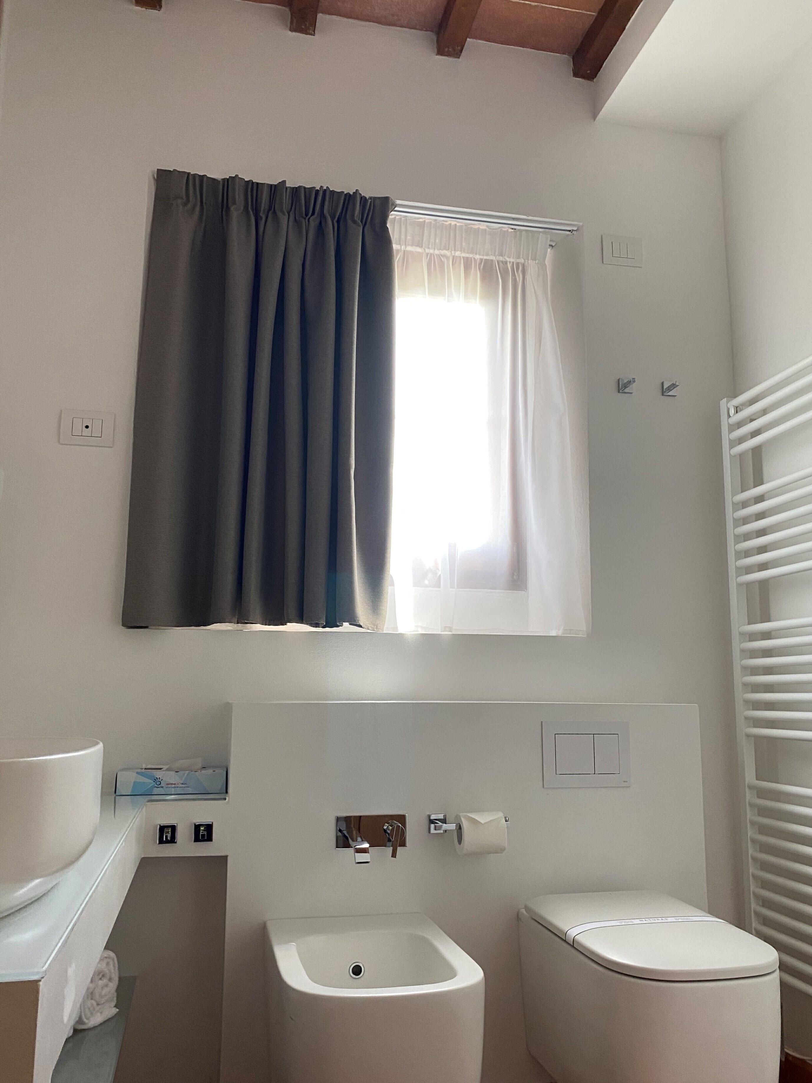 Baño con inodoro, bidé y lavamanos. Se ve una ventana con cortina, y un perchero para toallas.
